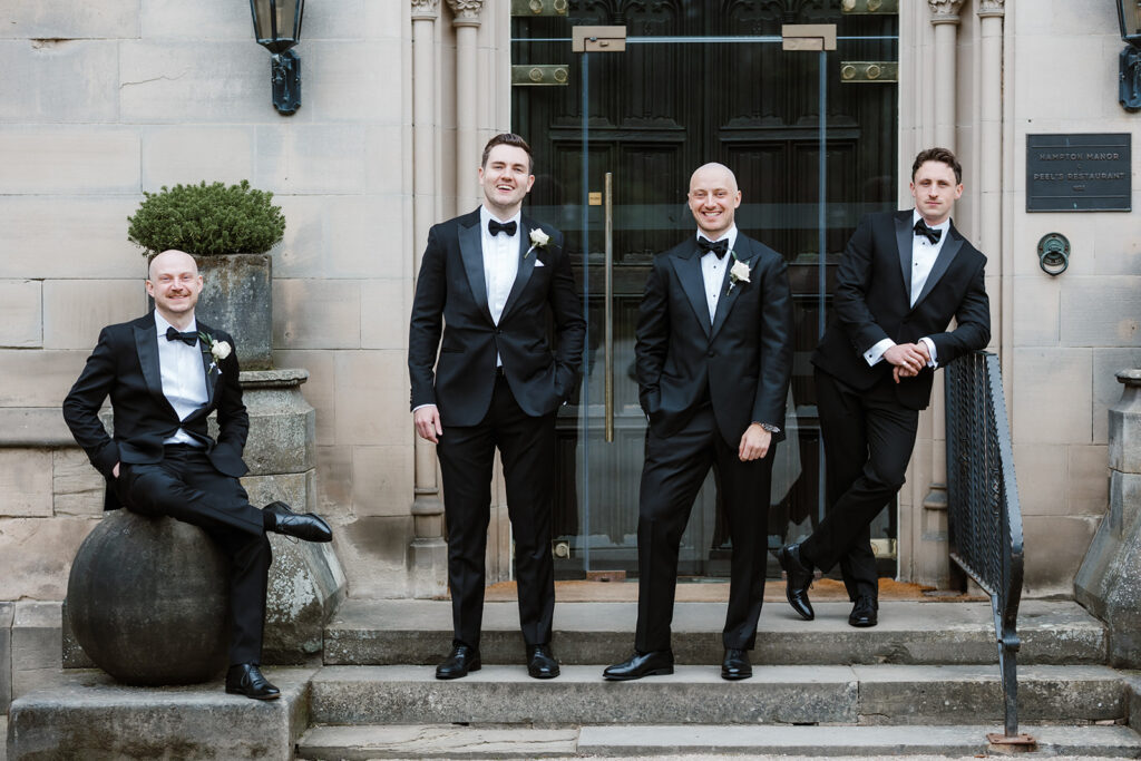 The groom and the groomsmen in dinner jackets pose for a formal photograph outside a stone building, standing and leaning casually on the steps and against a planter.