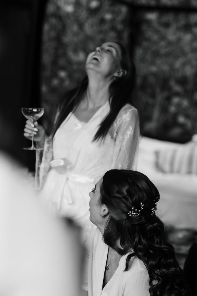 Two women in dressing gowns; one sitting and one standing with a drink, both appearing to laugh. The background is blurred. Image in black and white.