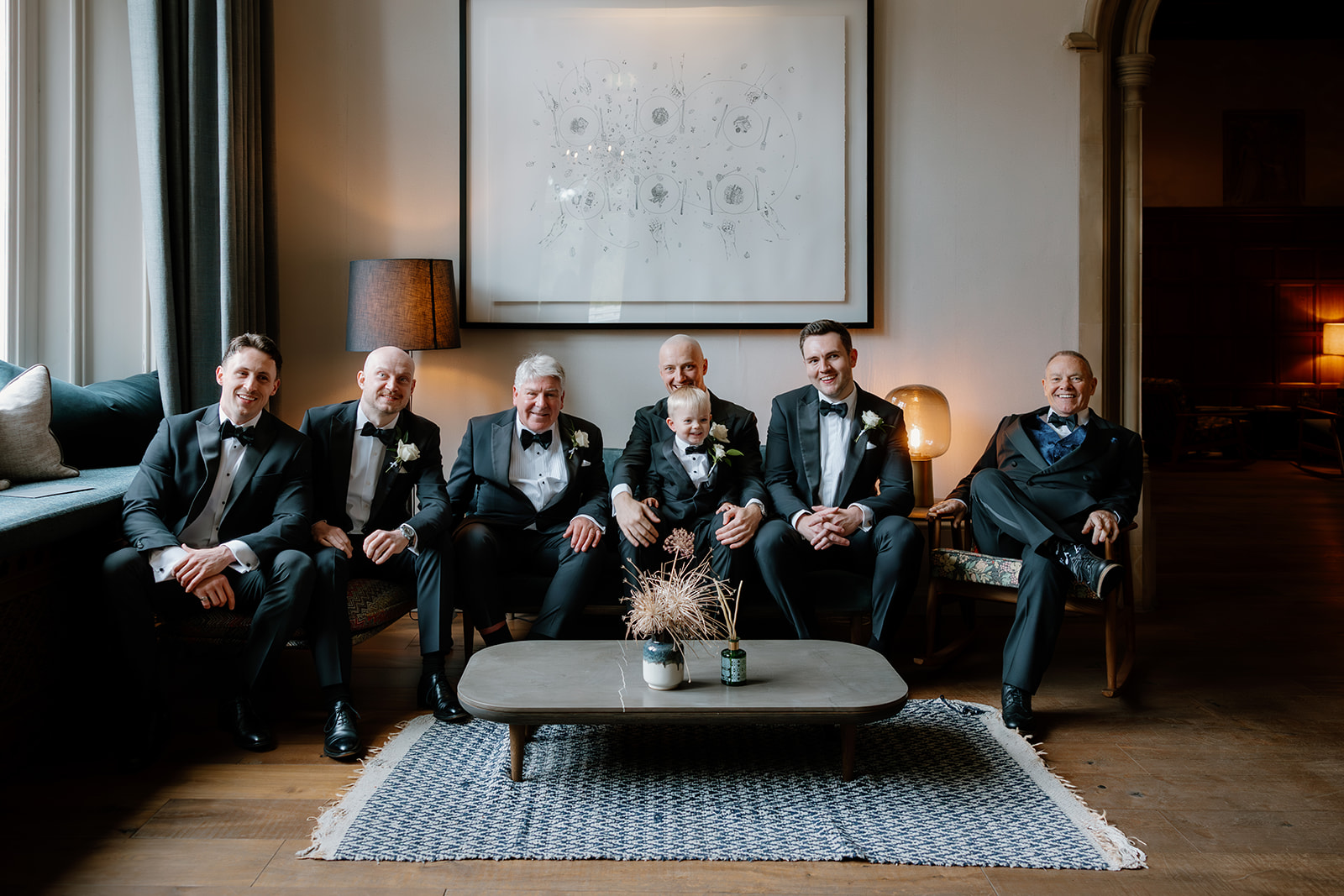 Six groomsman in formal suits sit on a sofa and chairs in a well-lit sitting room with framed wall art behind them at Hampton Manor