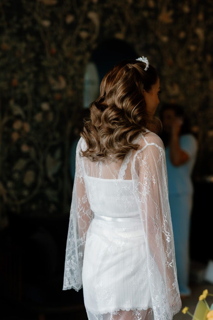 The bride with wavy hair, wearing a sheer lace dressing gown over a white outfit, stands indoors with her back to the camera.