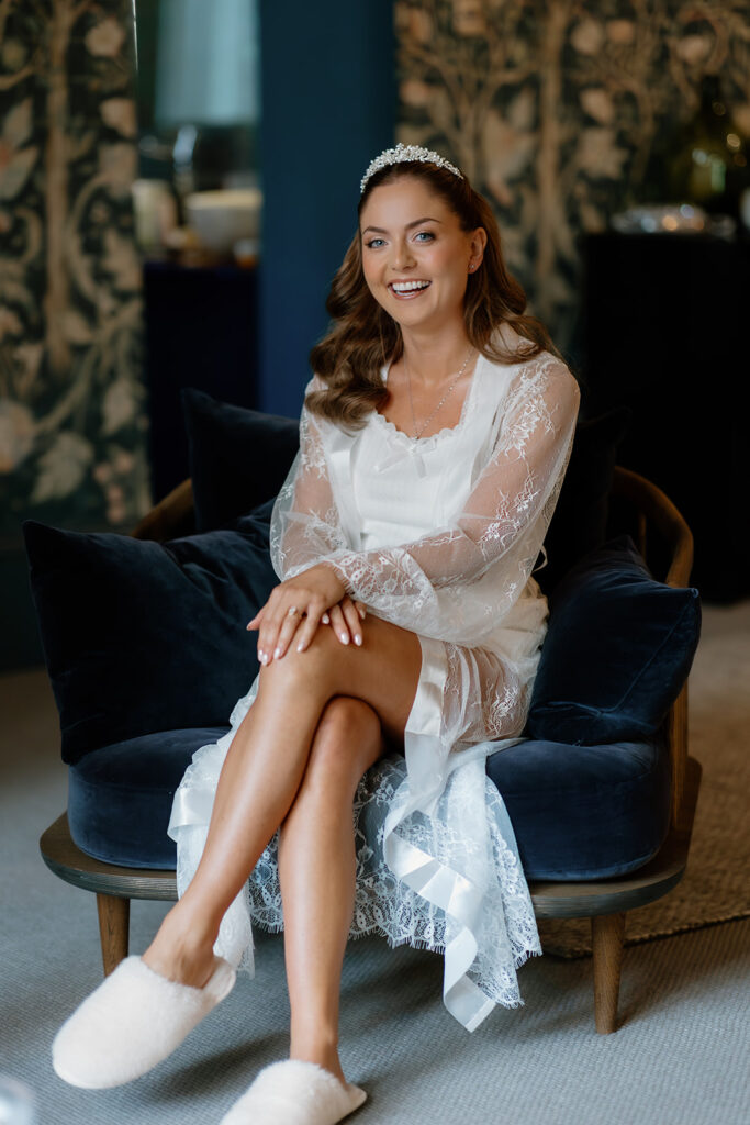 A bride in a white lace dressing gown and tiara sits on a dark armchair, smiling with her hands on her knee. She is wearing slippers and appears to be getting ready, possibly for a special occasion.