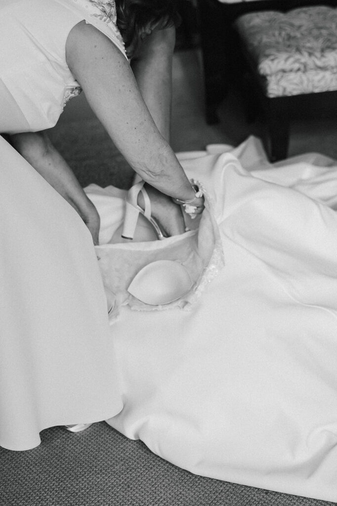 A mum helping her daughter step into her dress adjusts a pair of high-heeled shoes inside a basket on the train of a white dress, with part of a patterned chair visible in the background.