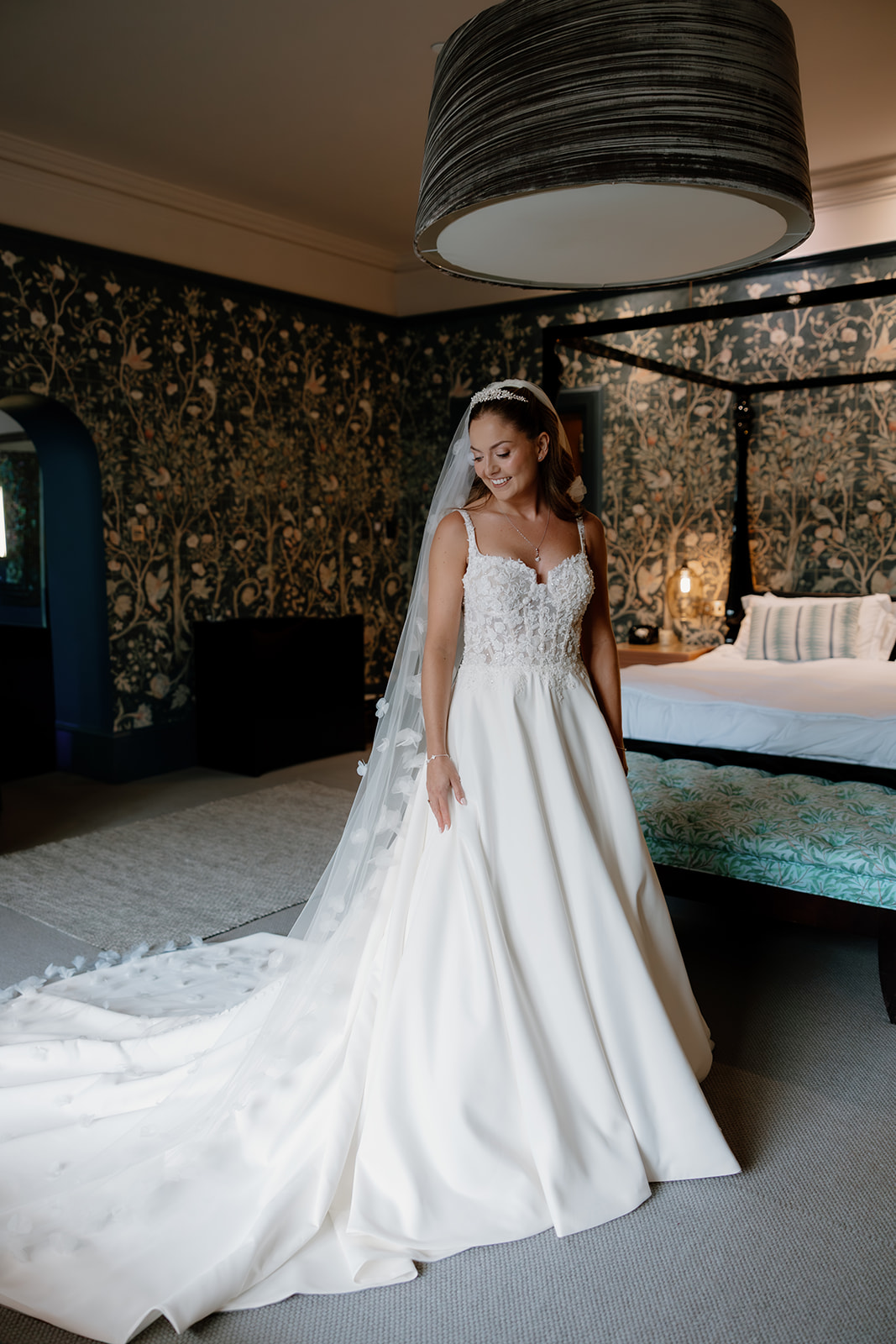 A bride in a white wedding dress with a long train and veil stands smiling in a decorated bedroom with patterned wallpaper.