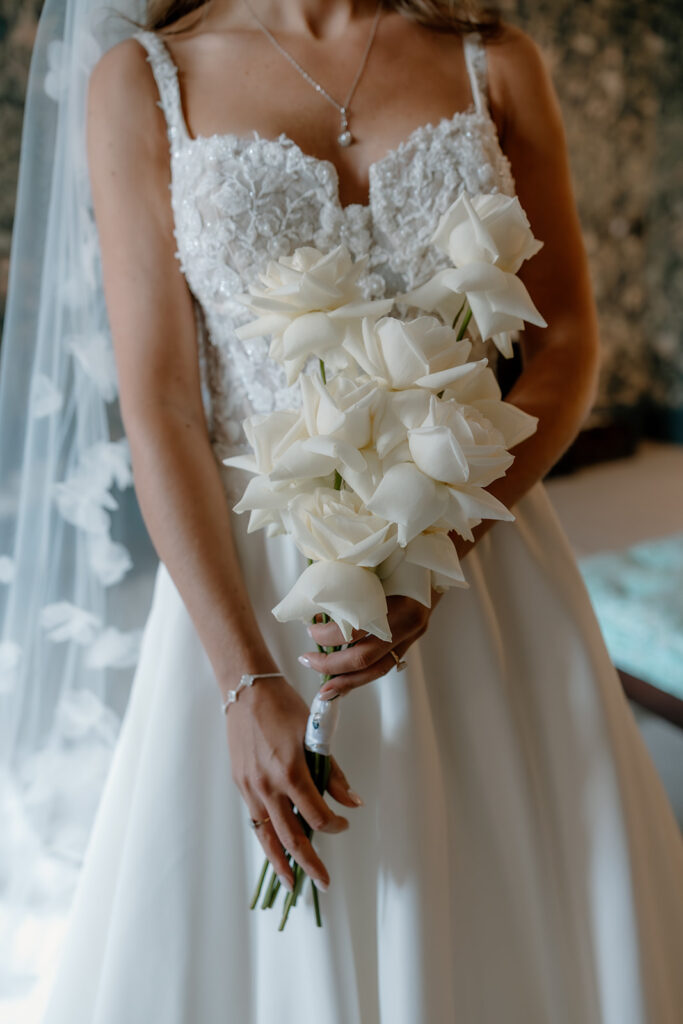 A bride in a white lace wedding dress holds a bouquet of white roses.