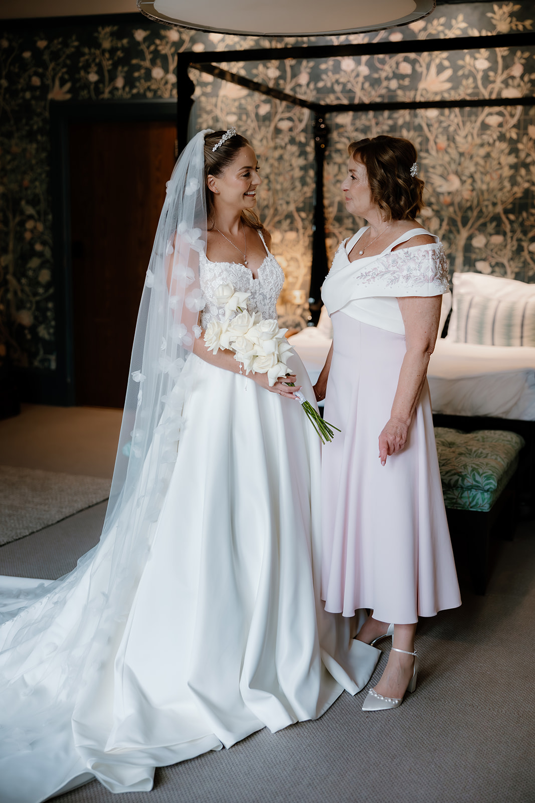 A bride in a white gown and veil holds a bouquet and stands beside a woman in a light pink dress, both smiling at each other in a bedroom.