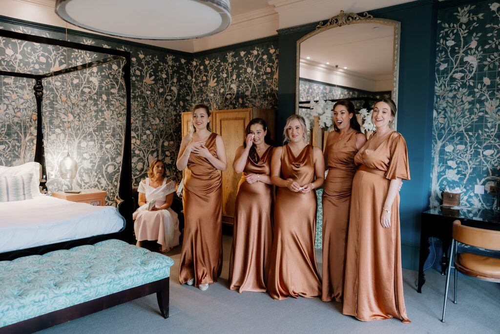 Five bridesmaids in matching bronze dresses stand together in a decorated bedroom, their first look reactions directed off-frame. In the background, a woman in white sits on a chair, watching the moment unfold.