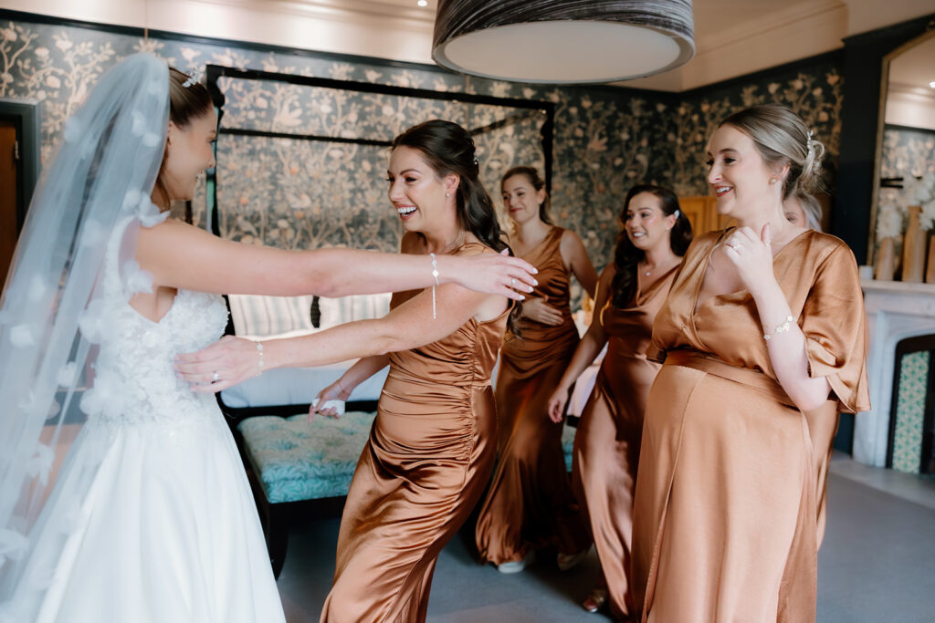A bride in a white dress hugs a smiling bridesmaid in a bronze gown, with three other bridesmaids in matching dresses watching in a decorated room.