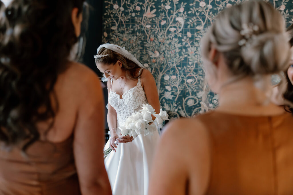 A bride in a white dress holding a bouquet of white flowers stands in front of a floral-patterned wall, with two women in brown dresses in the foreground.