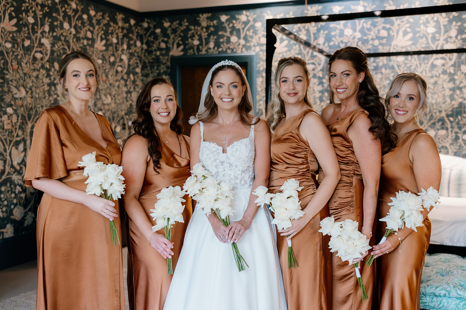 Five bridesmaids and the bride stand together indoors; one in a white bridal dress, the others in matching bronze dresses, all holding white flower bouquets and smiling at the camera.