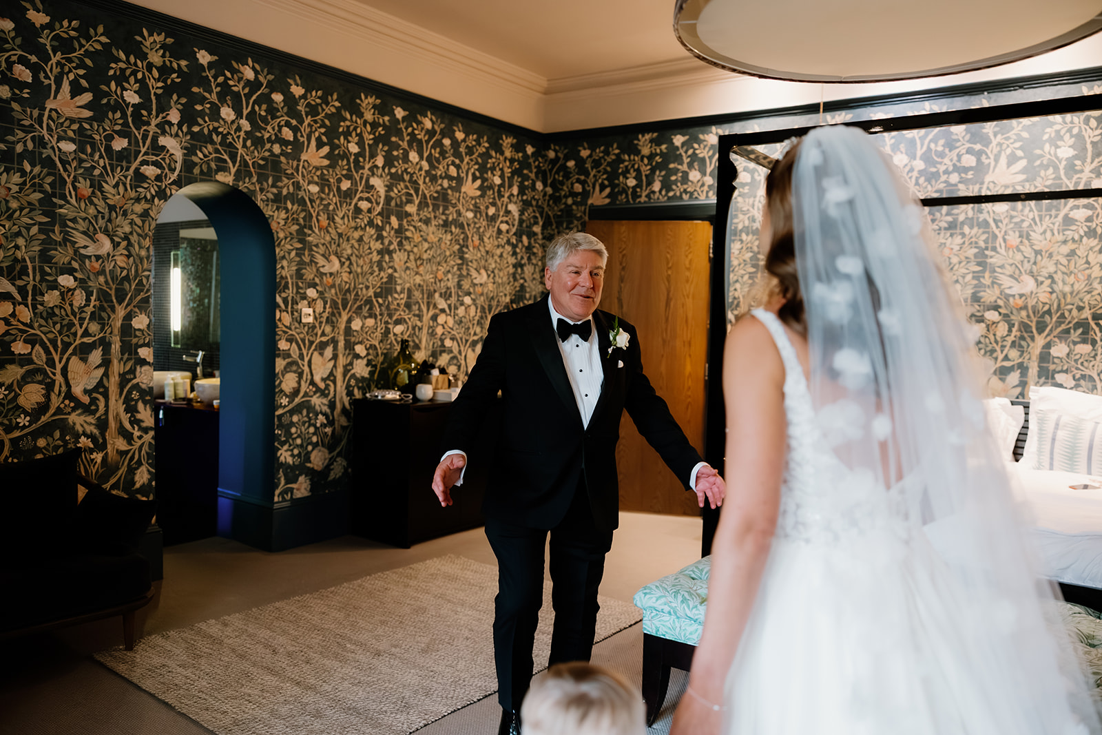 A father of the bride in a tuxedo stands with open arms, beaming during the emotional first look at his daughter in her white wedding dress and veil inside a beautifully decorated room.
