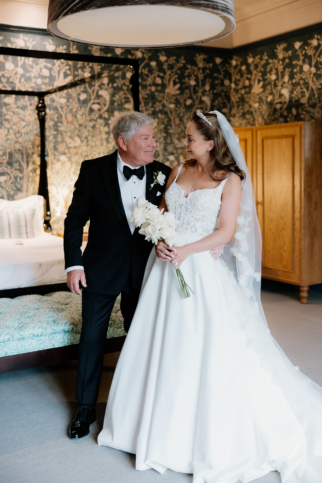 A bride and the father of the bride share a joyful moment in a decorated bedroom, smiling at each other as she stands in her white gown and veil, holding a bouquet beside him in his dinner jacket.