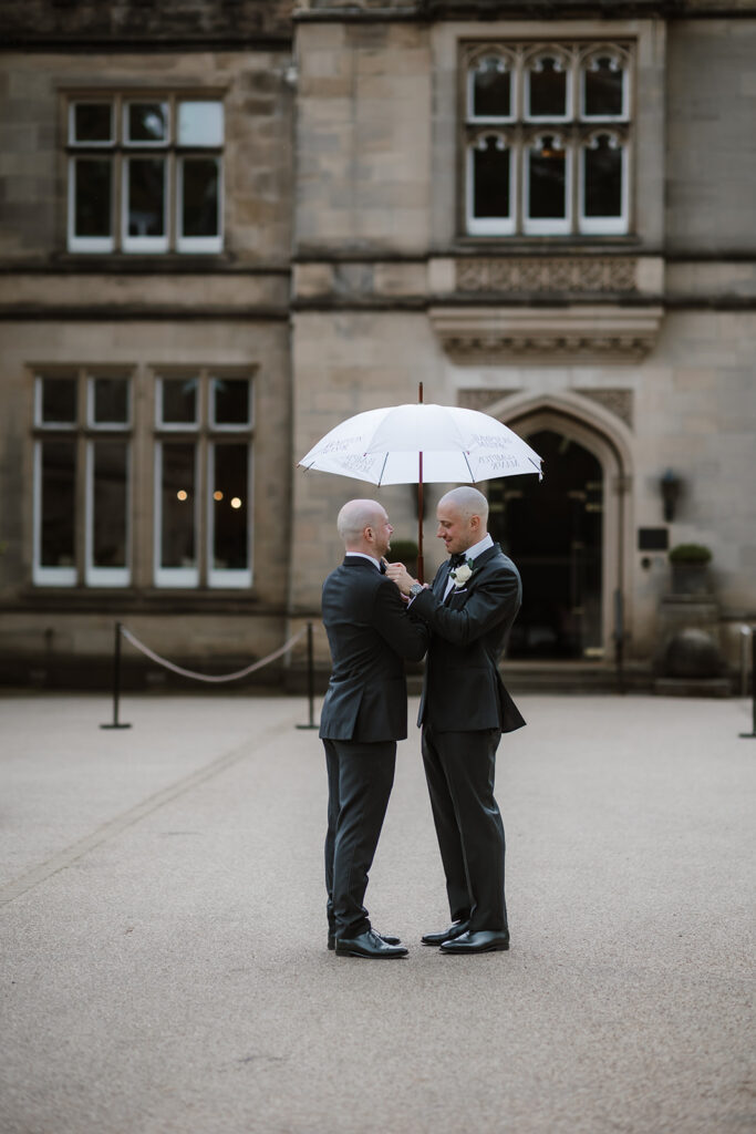 The groom and the best man, both in suits, stand facing each other under a white umbrella in front of a stone building, sharing a moment on a paved courtyard.