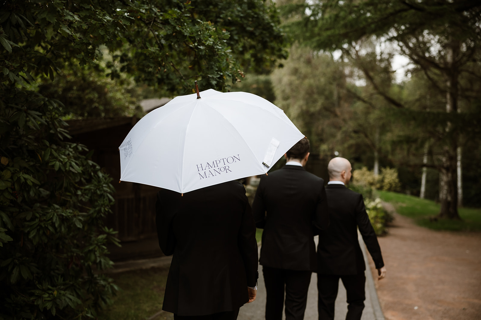 Three people in black suits walk on a path in a garden; one holds a white umbrella with "Hampton Manor" printed on it.