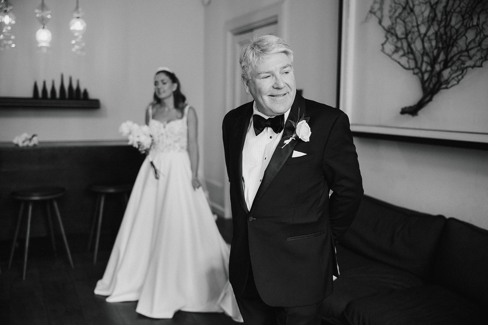 Father of the bride in a dinner jacket stands in the foreground, smiling, while a woman in a wedding dress stands in the background, both indoors.
