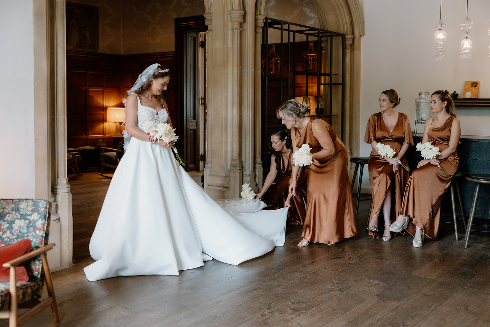 A bride at her Hampton Manor wedding stands in a white dress while four bridesmaids in bronze dresses sit and adjust her gown in an elegant room.