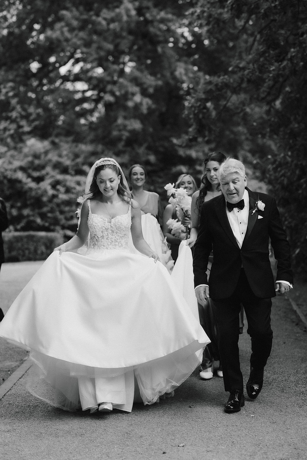 A bride in a wedding dress walks outdoors with several bridesmaids and an older man in a dinner jacket on a path surrounded by trees.