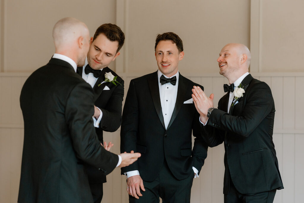 Four men in black dinner jackets stand together indoors, engaged in conversation, with two smiling and one applauding.