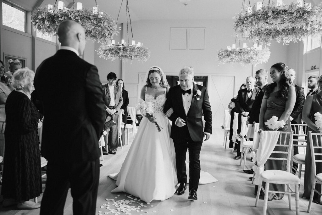 A bride in a wedding dress walks down the aisle with an older man, surrounded by standing guests in a decorated indoor venue.