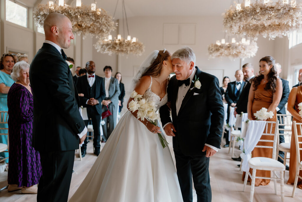 A bride in a white dress kisses an older man on the cheek whilst holding a bouquet, as guests and a man in a dinner suit look on in a decorated indoor wedding venue.