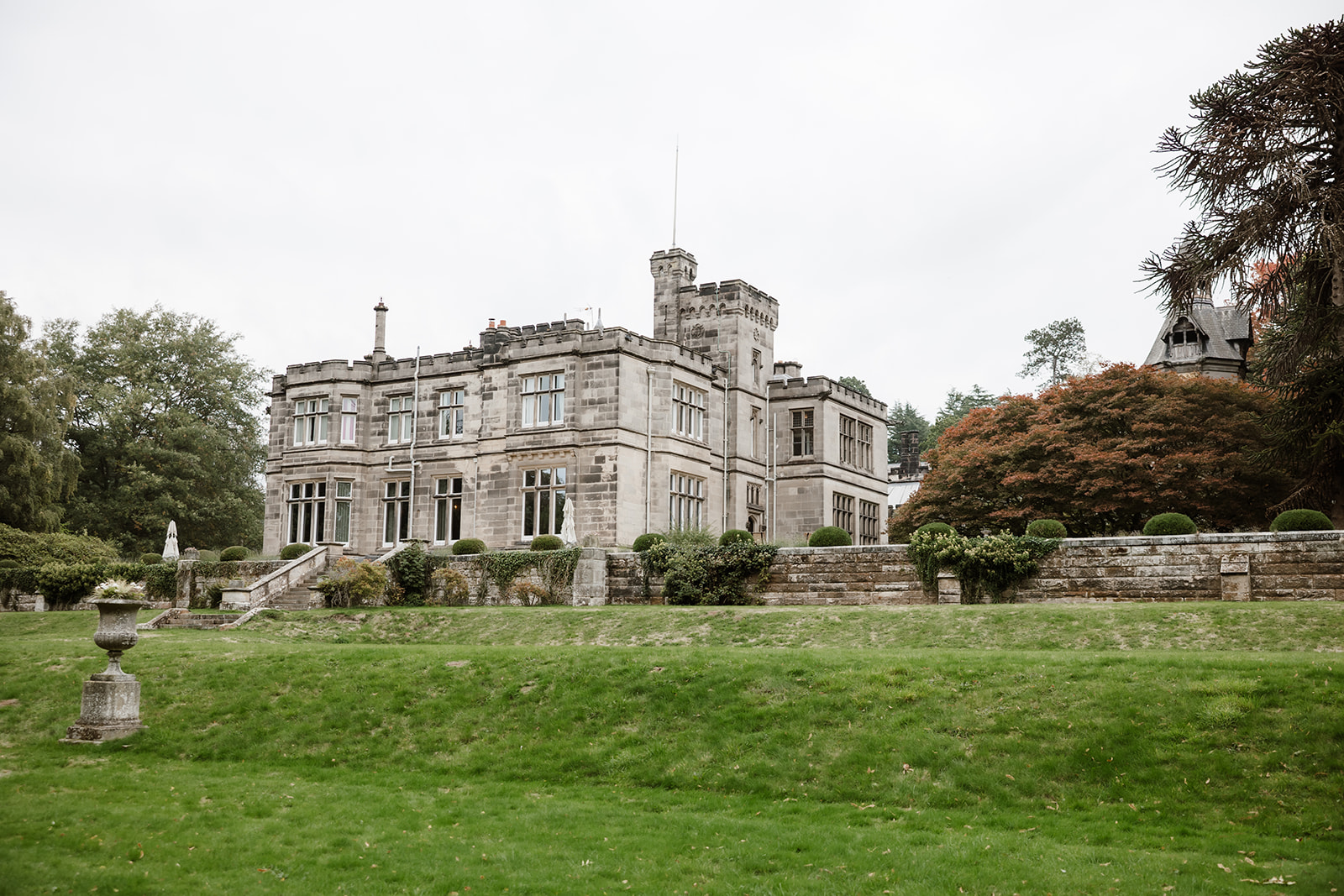 A large stone manor with multiple chimneys and tall windows sits on manicured lawns, surrounded by trees and shrubs under an overcast sky. Hampton Manor