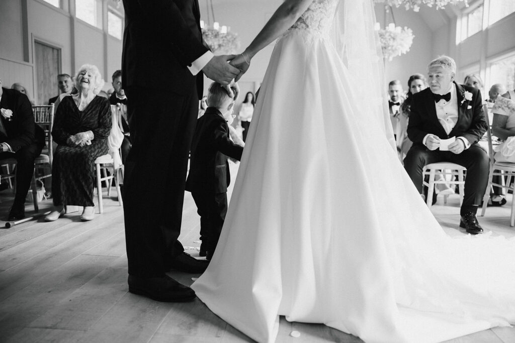 A bride and groom hold hands at their wedding ceremony as a young boy stands between them; guests are seated and watching.