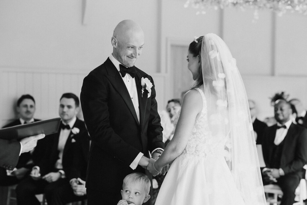 A bride and groom hold hands and smile at each other during their wedding ceremony, with a small child standing in front of them. Guests sit in the background.