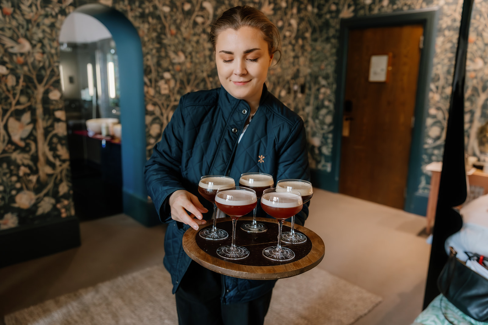 A person in a blue jacket holds a round tray with six espresso martini cocktails in a room with patterned wallpaper.