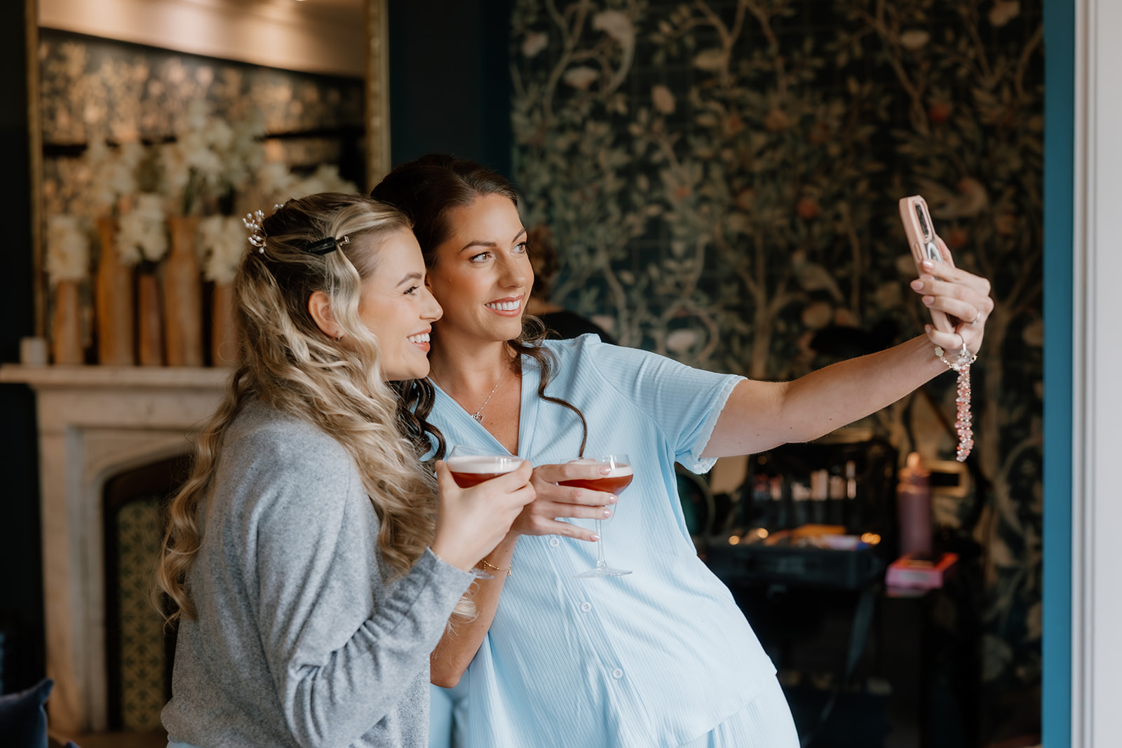 Two women smiling and holding drinks take a selfie together indoors, standing next to each other in front of a floral wallpapered wall.