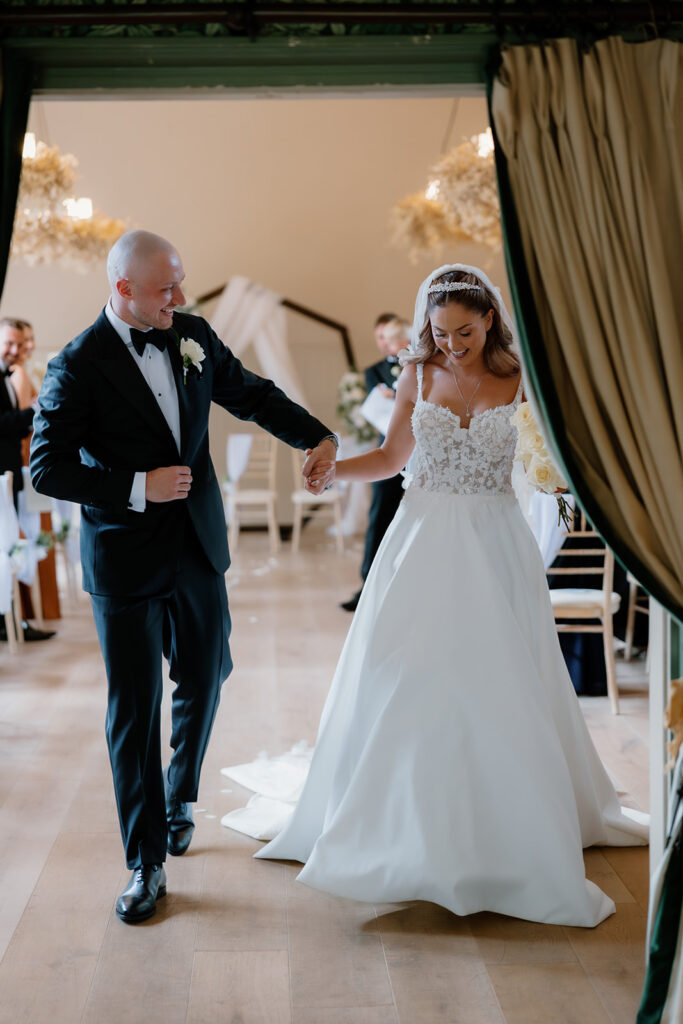 The happy bride and groom, holding hands and smiling, walk down the aisle together in a beautifully decorated indoor venue.
