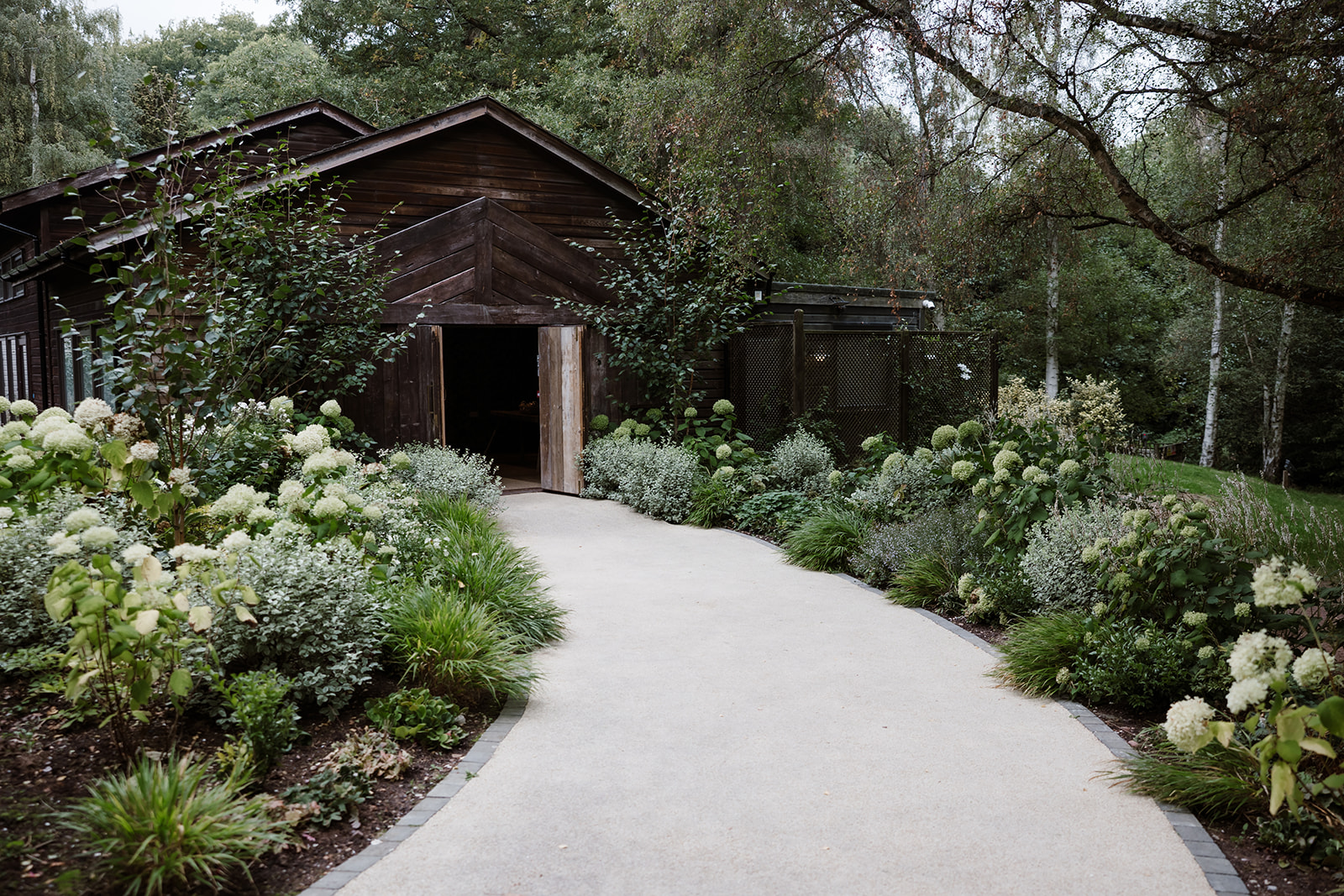 A paved path leads to a dark wooden building at Hampton Manor, surrounded by lush green plants and white flowers, with trees in the background. Birches ceremony room