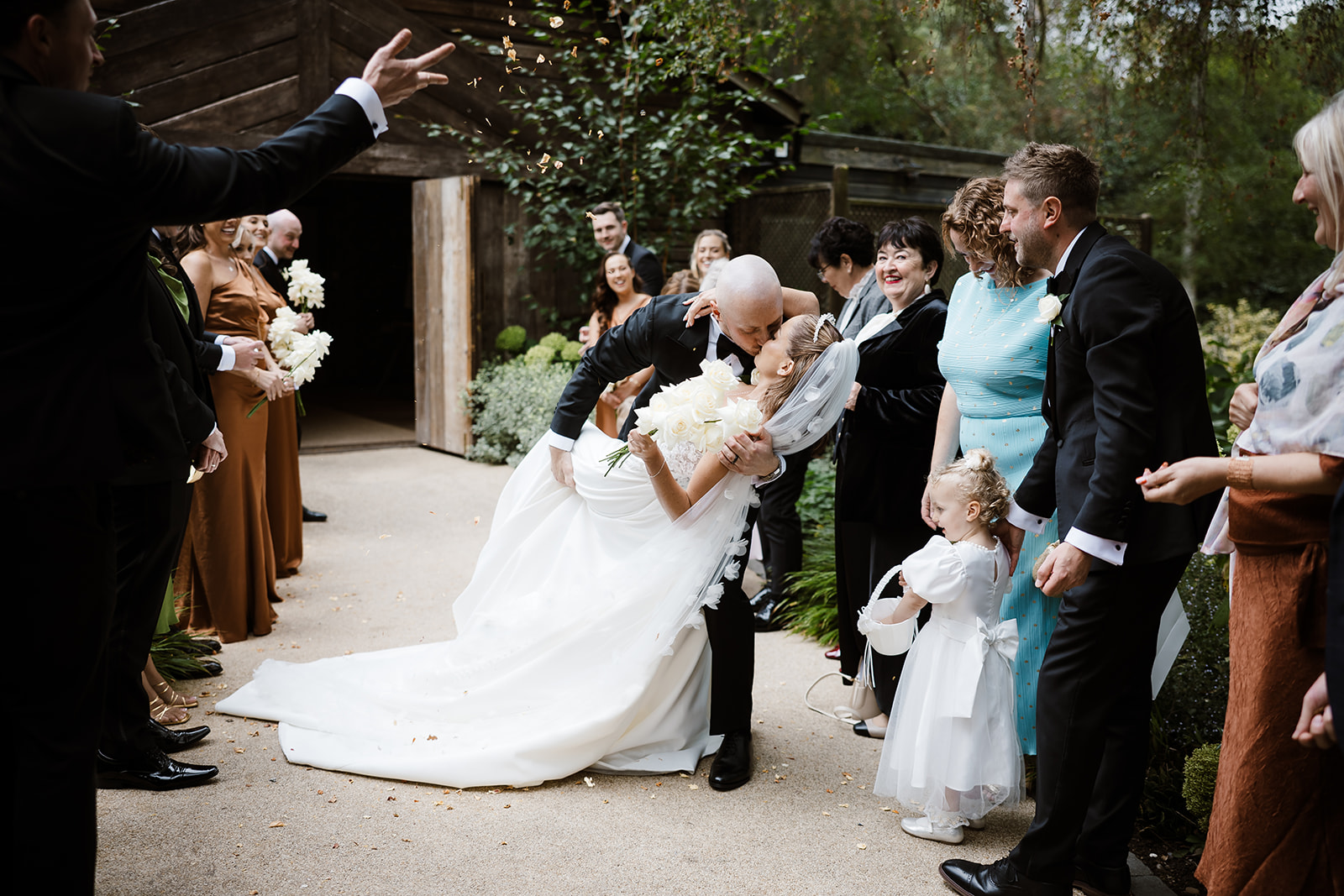 A newlywed couple kiss while guests, dressed in formal attire, stand around them outdoors at Hampton Manor, holding flowers and smiling. A perfect moment captured by a Hampton Manor wedding photographer.