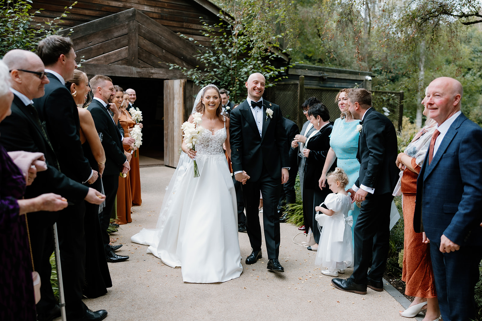 A bride and groom walk down an outdoor aisle, smiling and holding hands, surrounded by clapping and cheering guests.