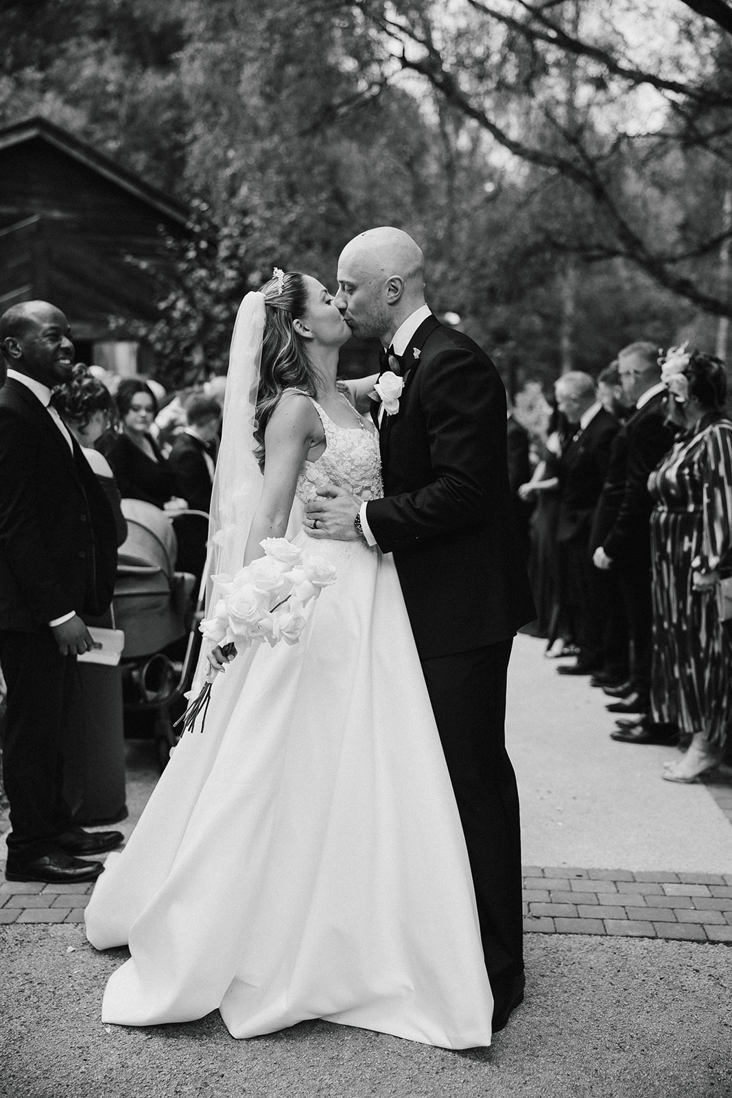 A bride and groom kiss outdoors during their wedding ceremony, surrounded by guests, in a black and white photograph.