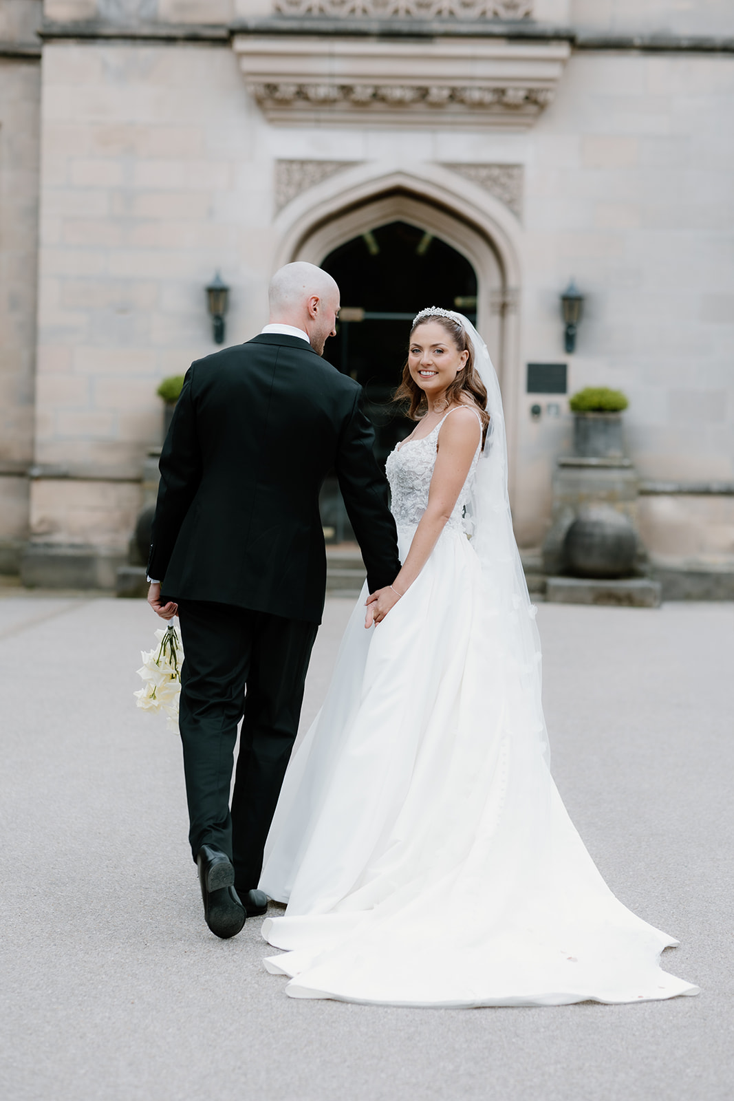 A bride and groom walk hand in hand outside a stone building; the bride wears a white dress and veil, while the groom holds a bunch of flowers and wears a black suit.