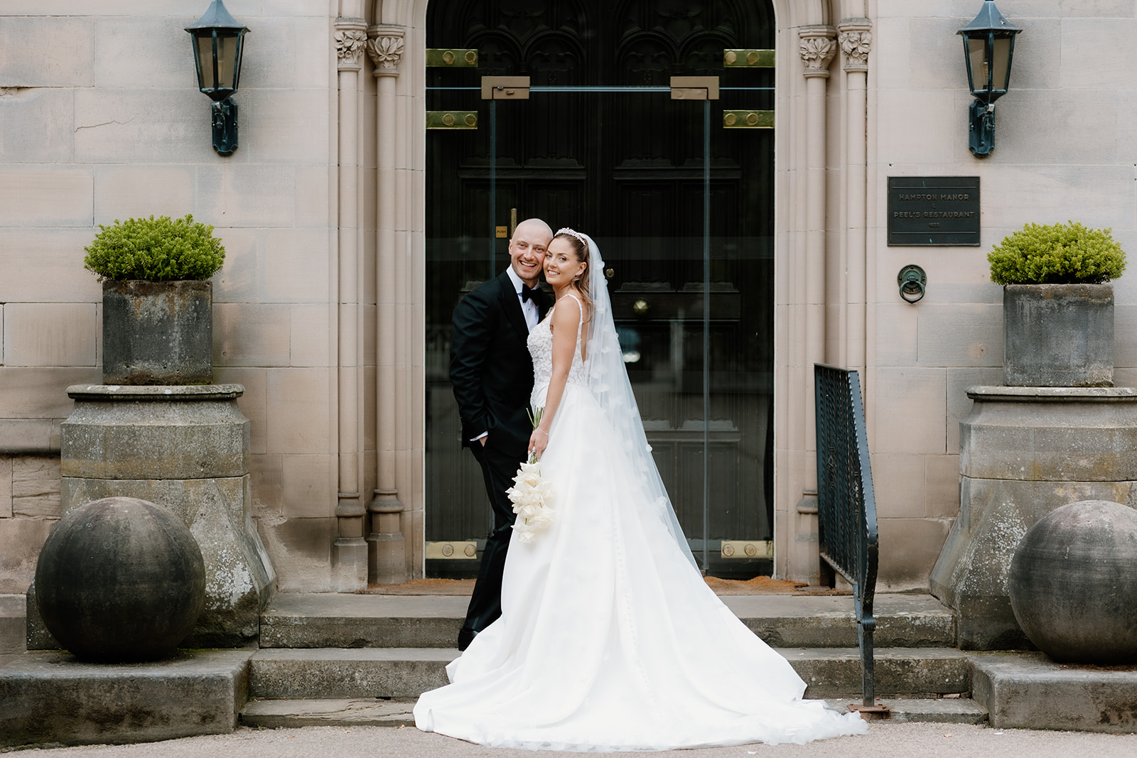 A bride and groom stand together on stone steps in front of a formal building, both smiling at the camera. The bride wears a white dress and veil, holding a bouquet.