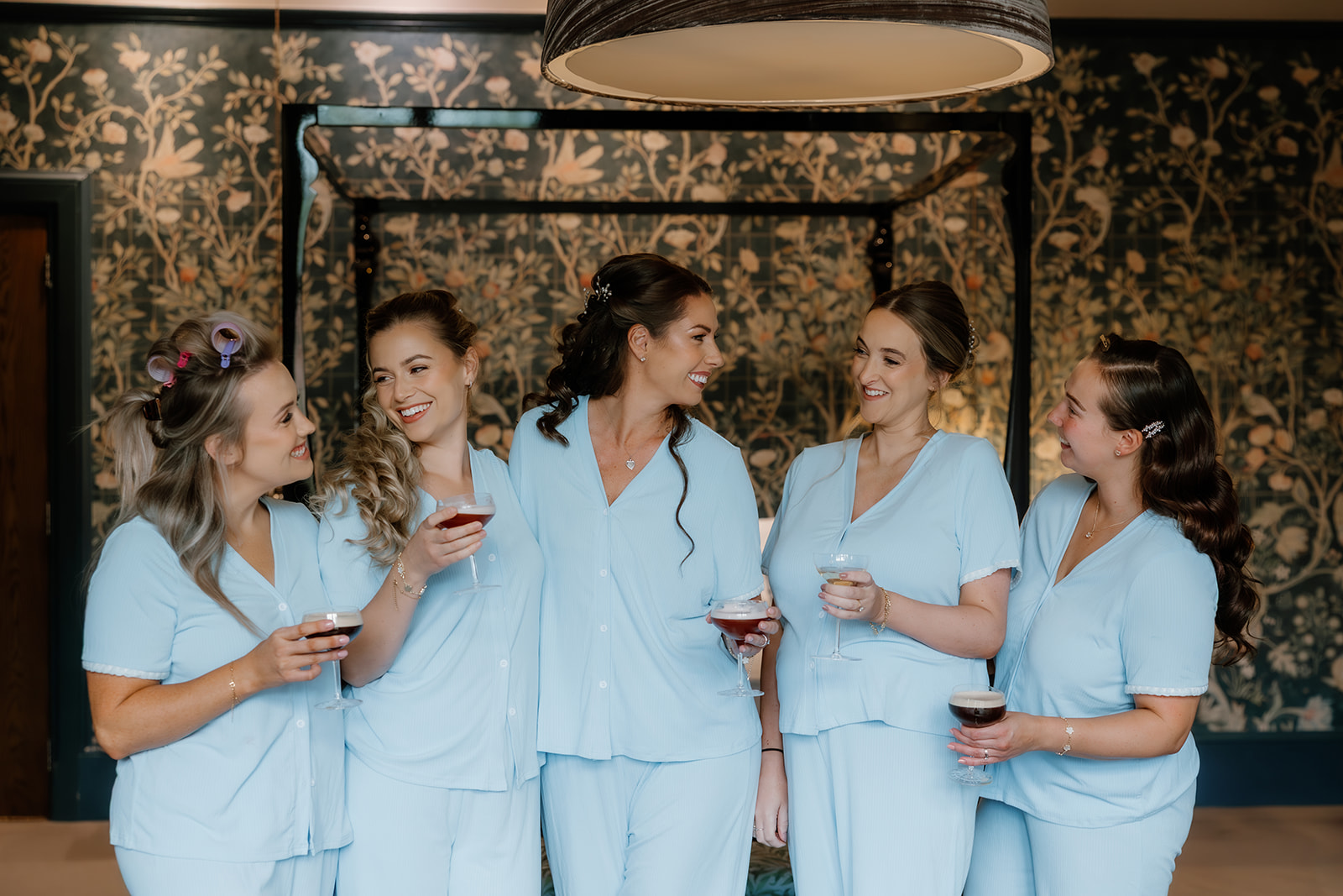Five bridesmaids in matching light blue pyjamas stand together indoors, holding drinks and smiling at each other in front of a decorative floral wallpaper background.