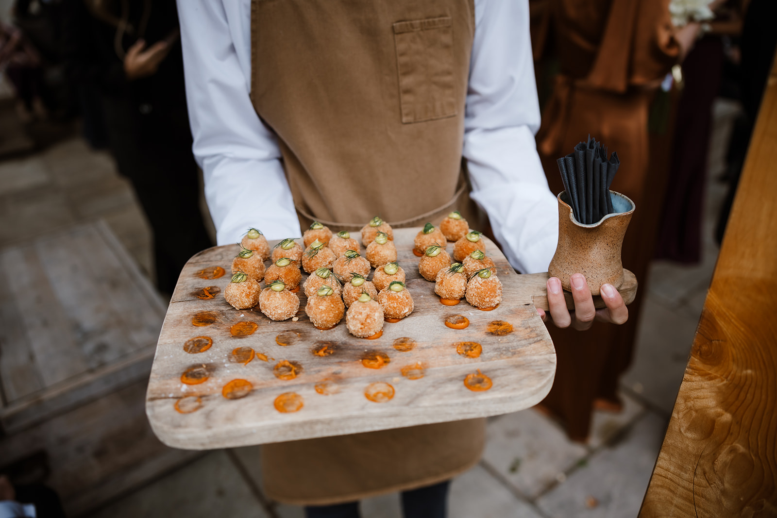 A person wearing a brown apron holds a wooden tray of round, breaded appetisers topped with herbs, with dots of orange sauce, and a cup of black cocktail straws.