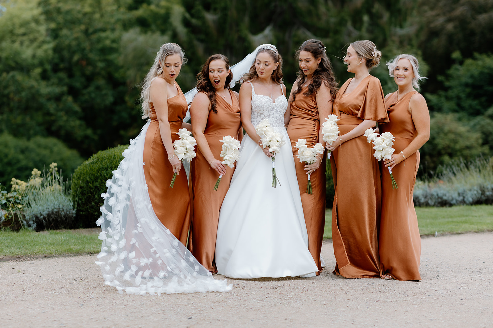 A bride in a white dress stands outside with her bridesmaids, all in matching burnt orange dresses and holding white flower bouquets.