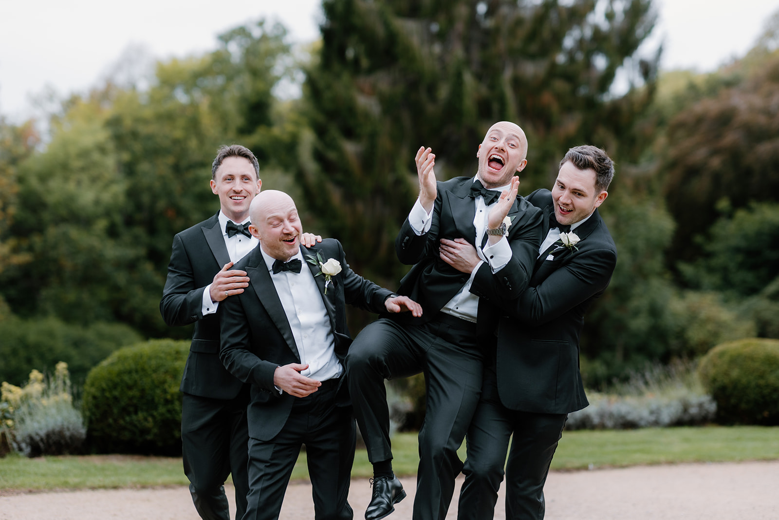 Four men in black dinner jackets, likely the groom and groomsmen, are laughing and posing playfully together outdoors, with greenery and shrubs in the background.