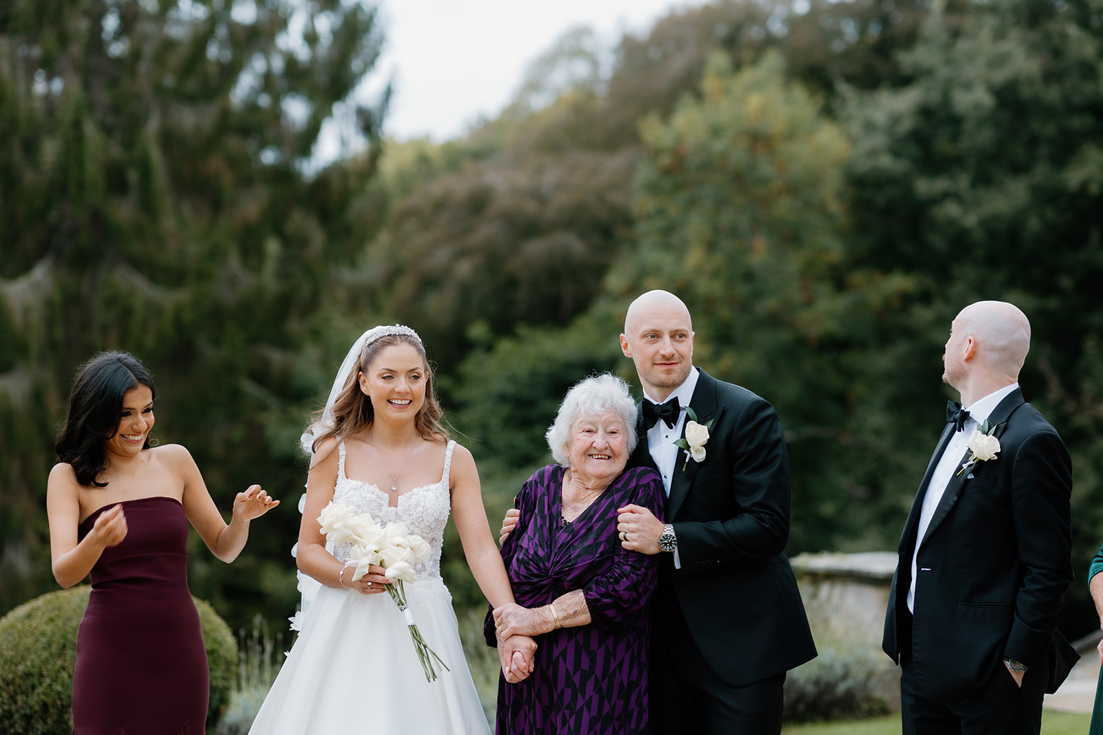 A bride, groom, and elderly woman stand together outdoors, smiling, with two other well-dressed guests nearby in a garden setting.