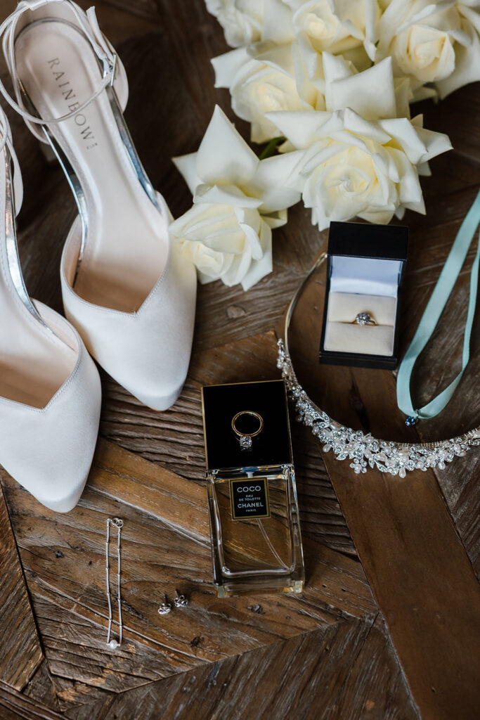 White high-heeled shoes, white roses, a diamond ring in a box, a jewelled headband, a perfume bottle, and jewellery pieces arranged on a wooden surface.