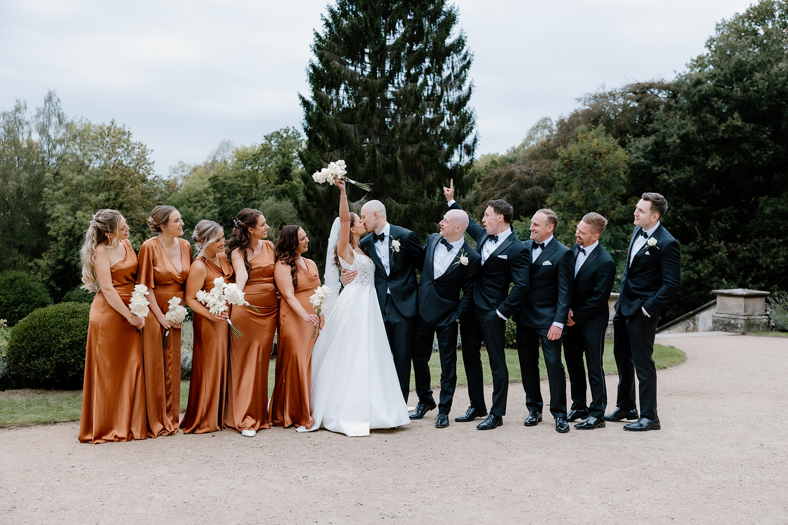 A joyful wedding party poses outdoors in front of trees; the bride and groom are surrounded by bridesmaids in matching rust dresses and groomsmen in black suits, all celebrating together.