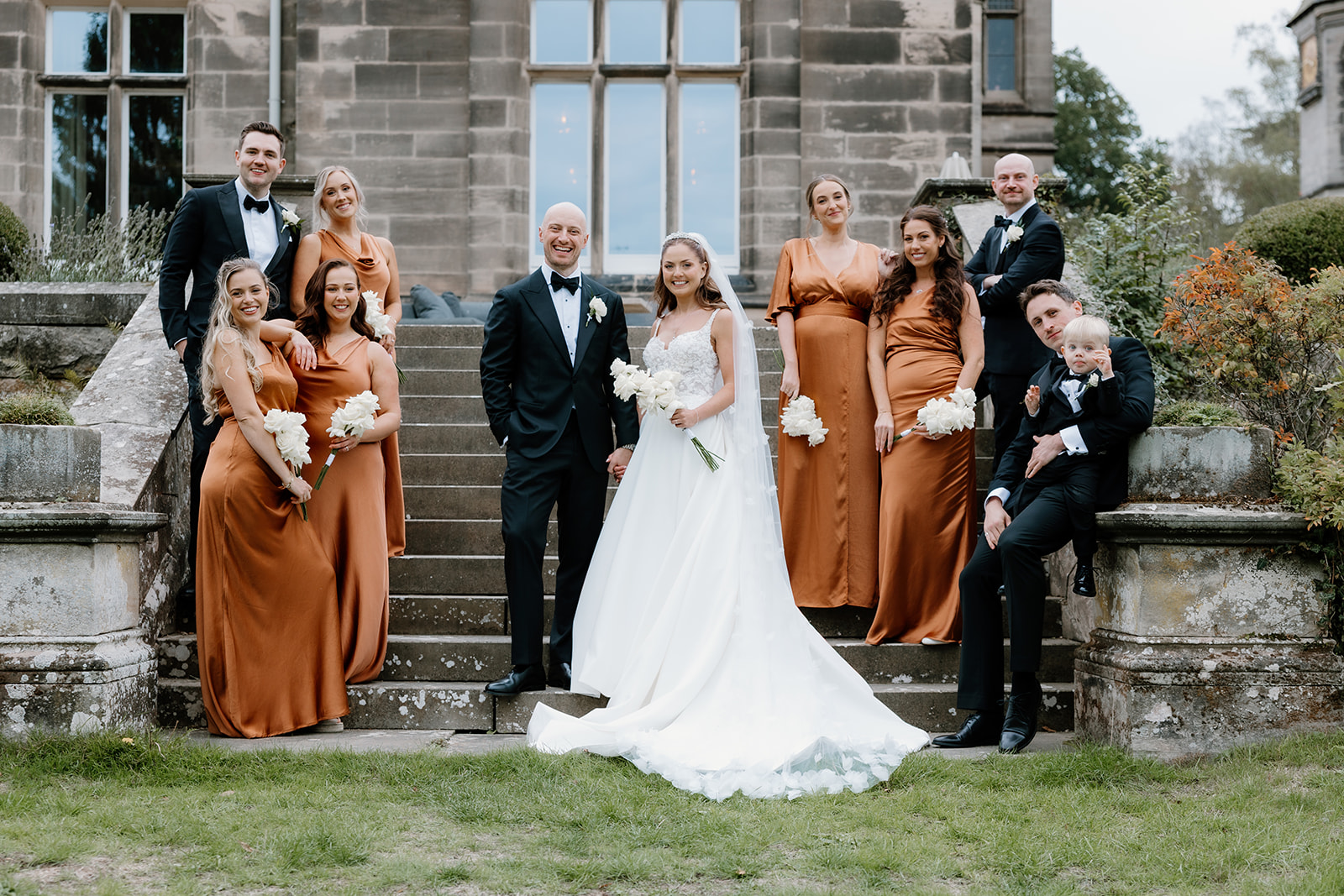 A wedding party poses on stone steps outside a building, with the bride and groom surrounded by bridesmaids in rust-coloured dresses and groomsmen in black suits.