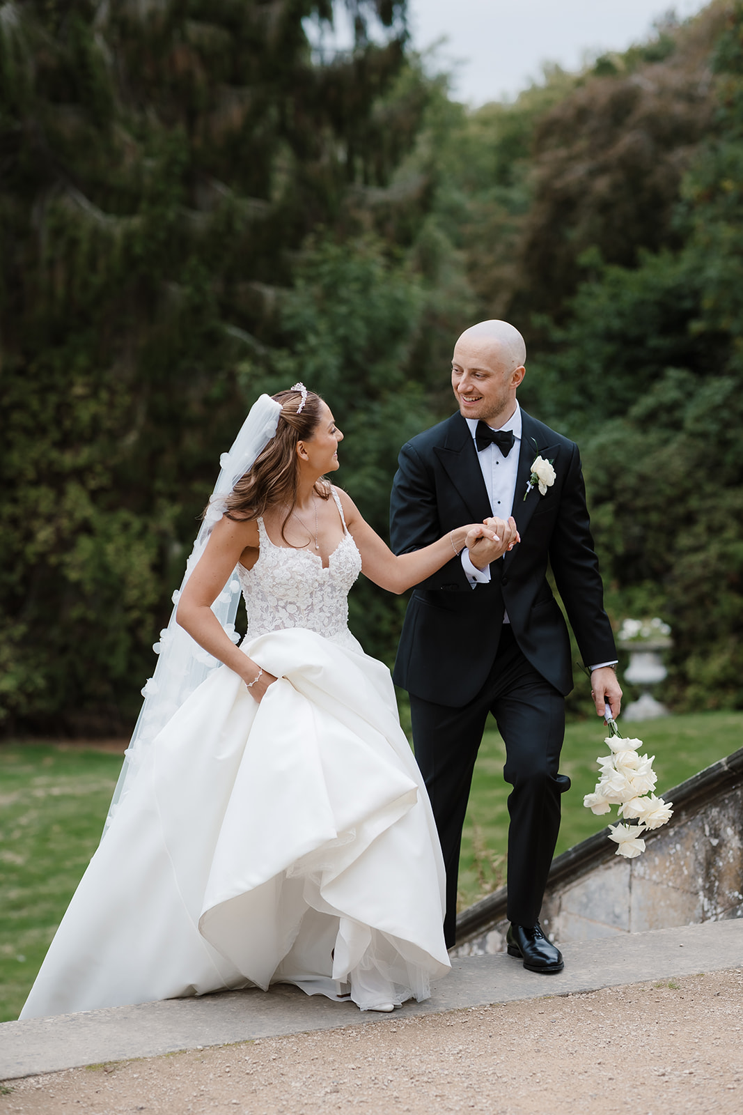 A bride in a white gown and a groom in a black tuxedo walk hand in hand outdoors at their Hamton Manor wedding, smiling at each other with lush greenery in the background.