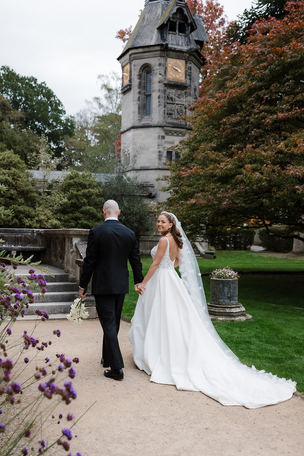 A bride and groom walk hand in hand outdoors near a historic stone tower, surrounded by greenery and flowers.