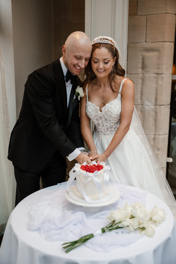 A bride and groom cut their elegant wedding cake adorned with strawberries, whilst a bouquet of white flowers sits gracefully on the table beside them.