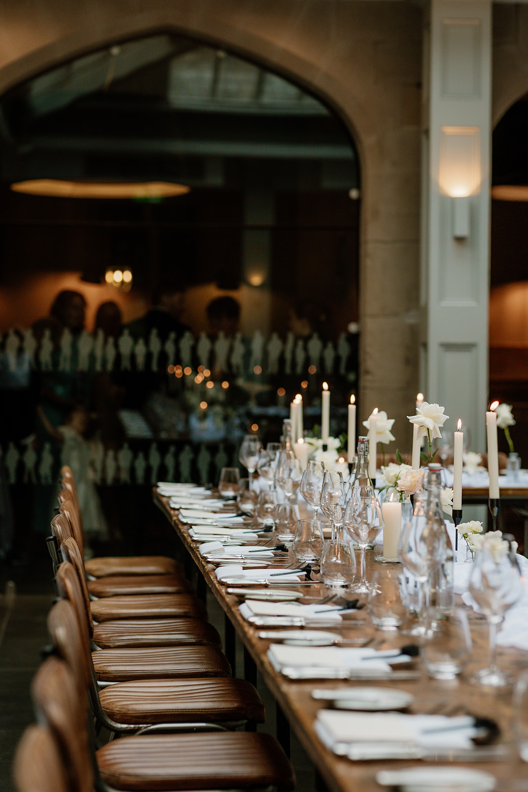 A long dining table set with plates, glasses, cutlery, white candles, and flowers in a softly lit room; people are visible in the background.