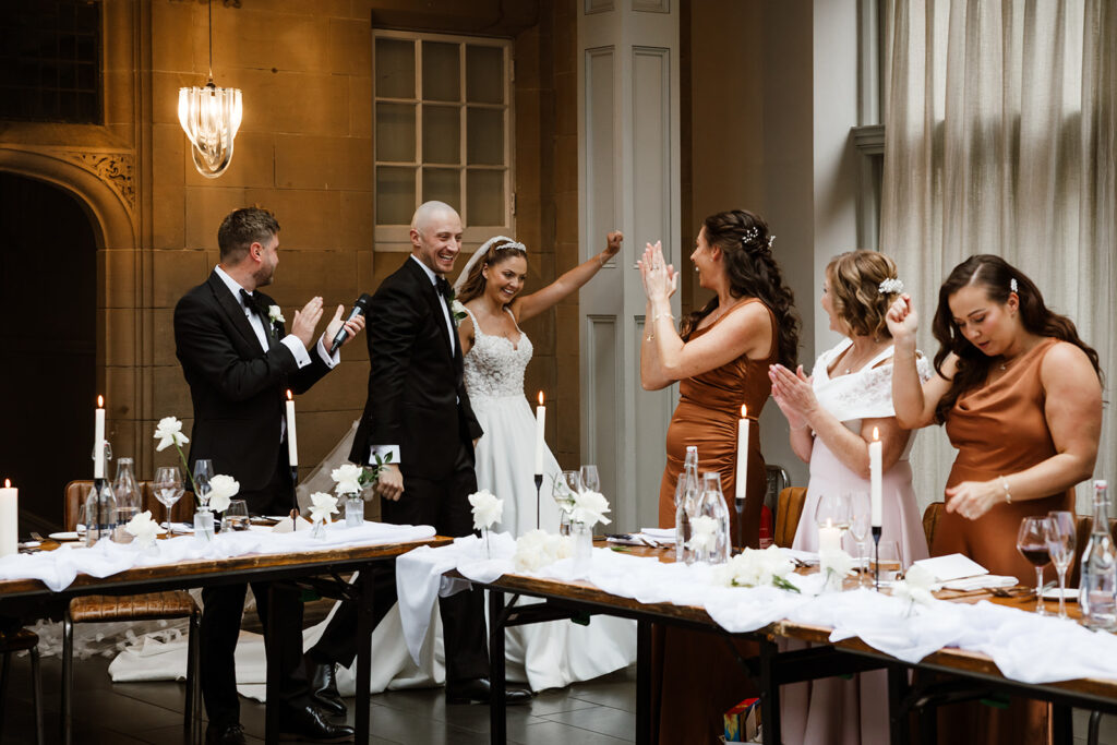 A bride and groom celebrate at their wedding reception while guests in formal attire applaud around a decorated table.