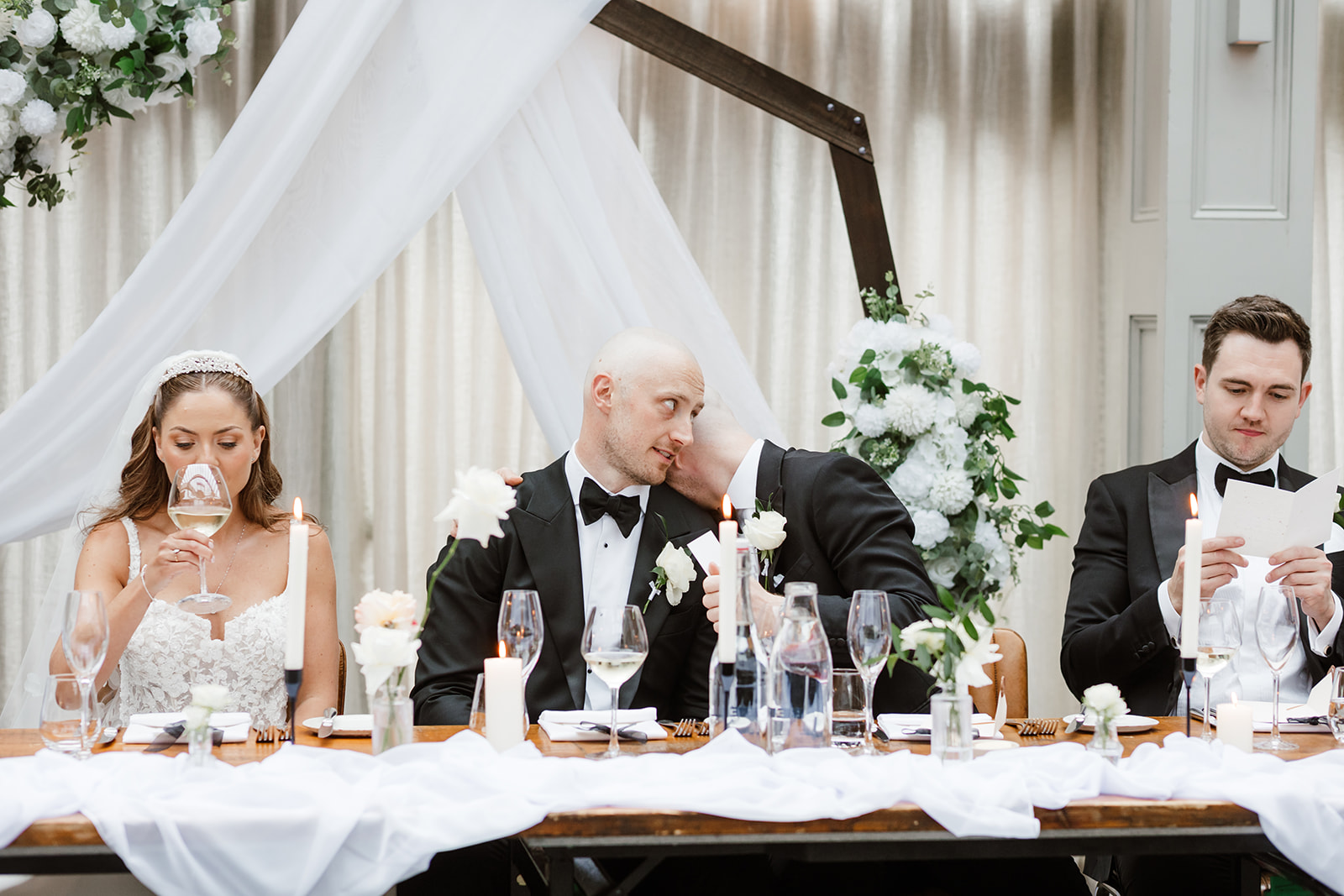 Four people in formal attire sit at a decorated wedding table; one man whispers to another, while a woman drinks wine and another man reads a piece of paper.