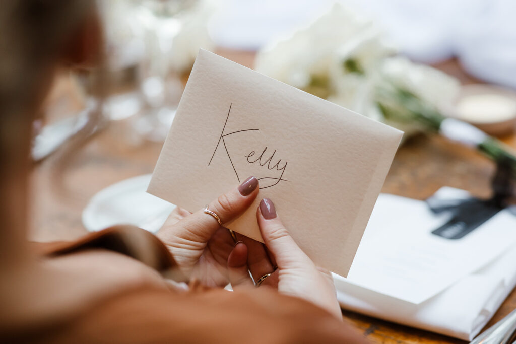 A person holding an envelope with the name "Kelly" written on it in cursive, with a blurred bouquet and papers in the background.
